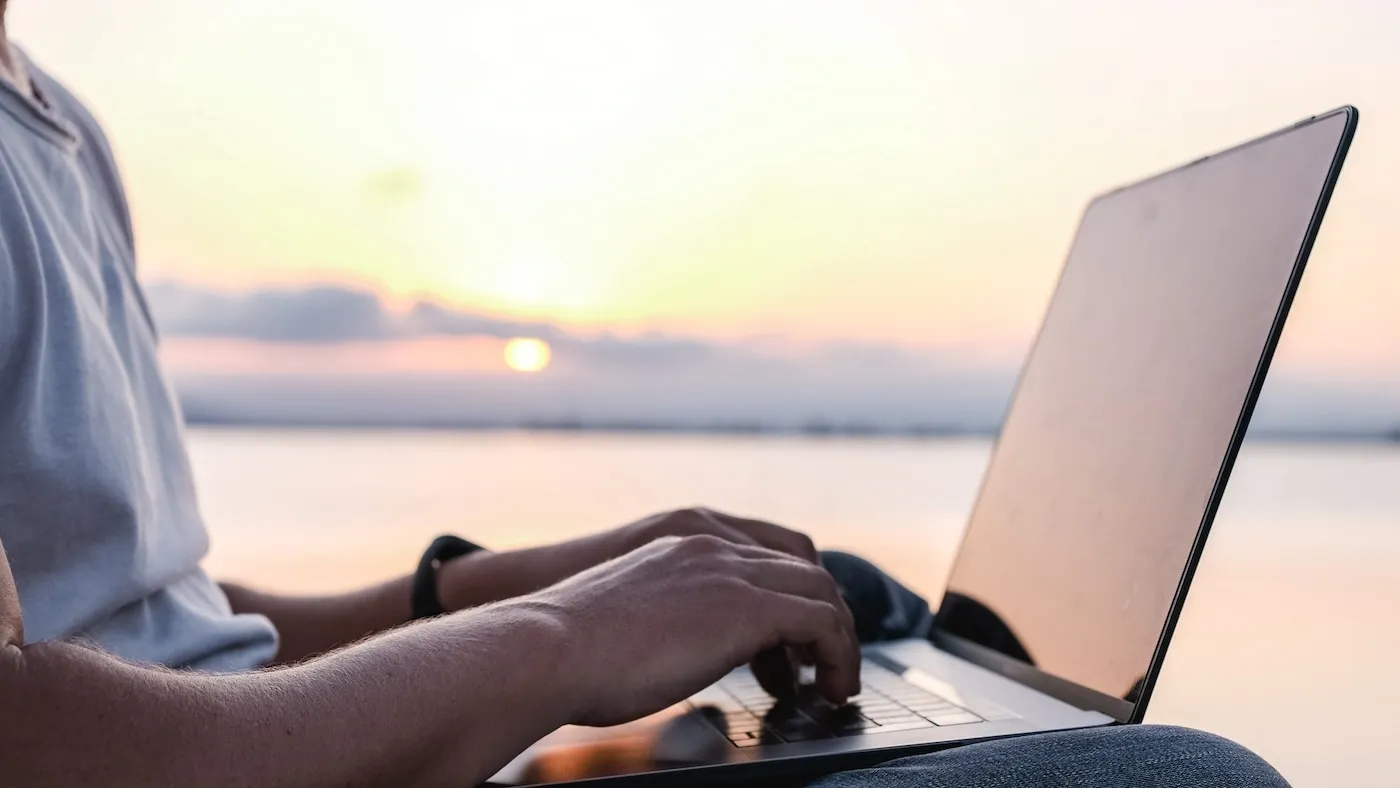 Person holding laptop with sunset in the background, at the beach.